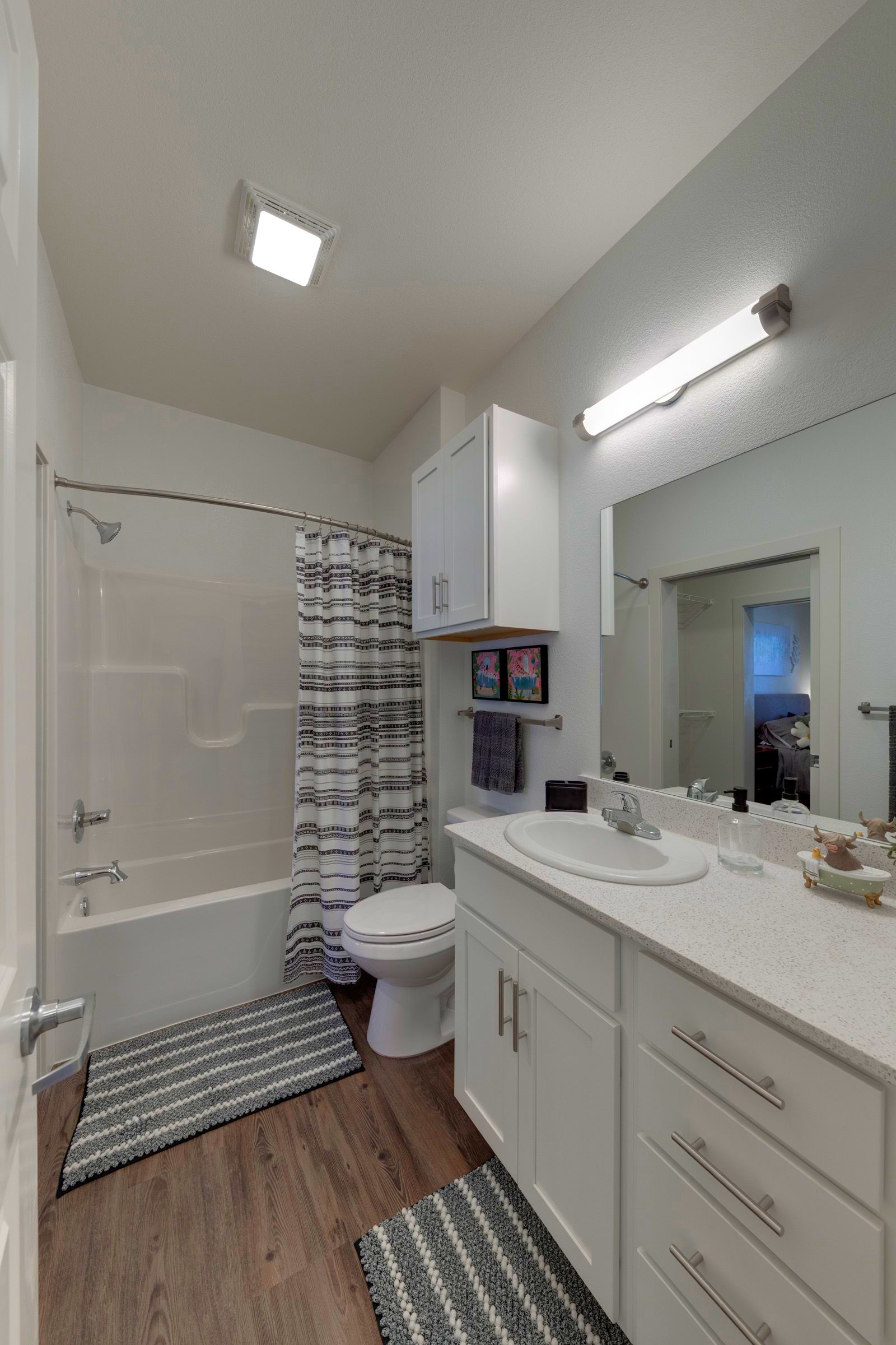 Bathroom with white cabinets, toilet, and tub; wood-look floor and gray striped rug.
