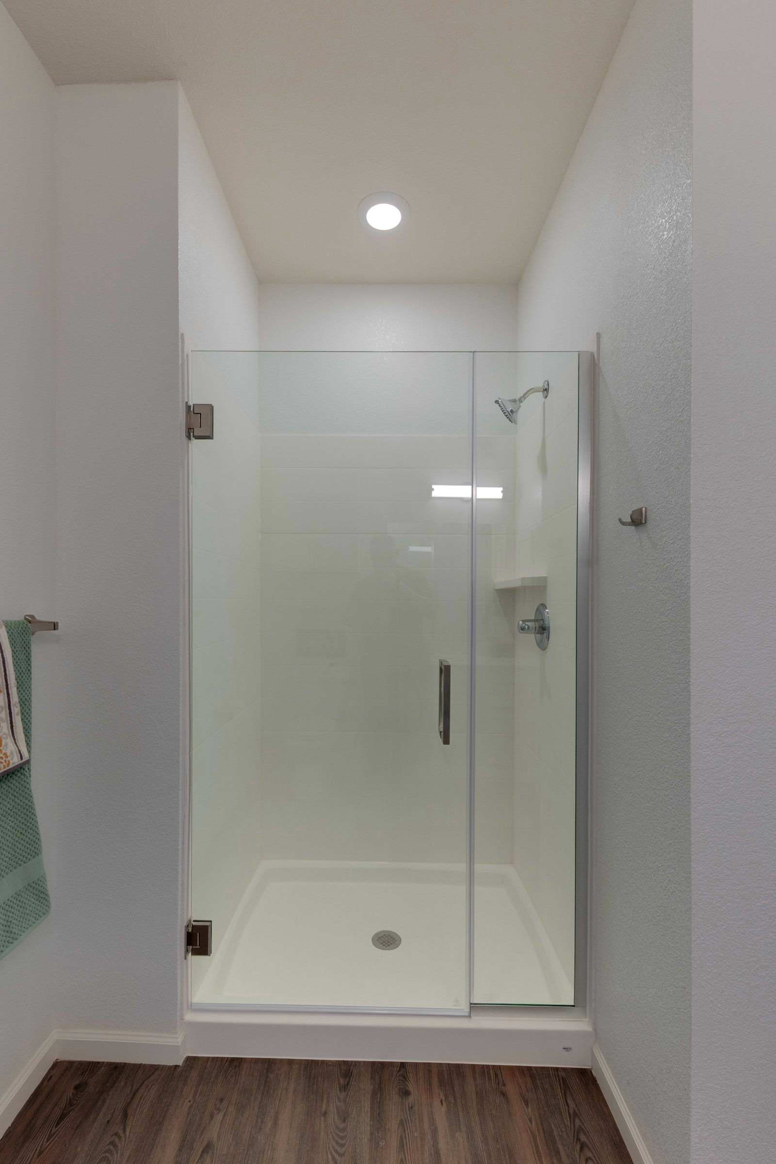 A small, white-walled shower stall with glass door and white basin, inside a narrow bathroom.