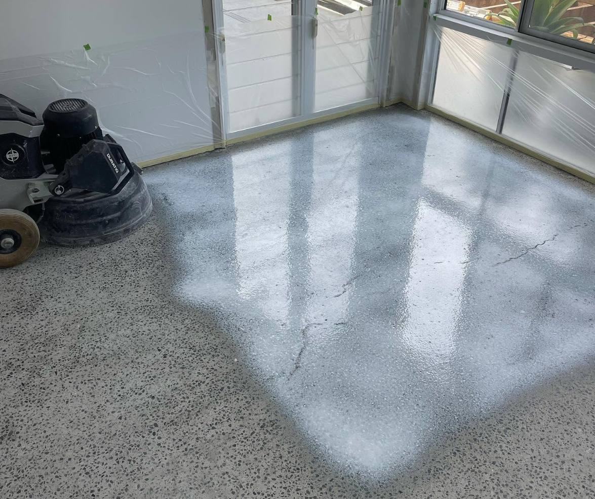 Polished speckled floor in a sunlit room with reflections and a vacuum cleaner in the corner