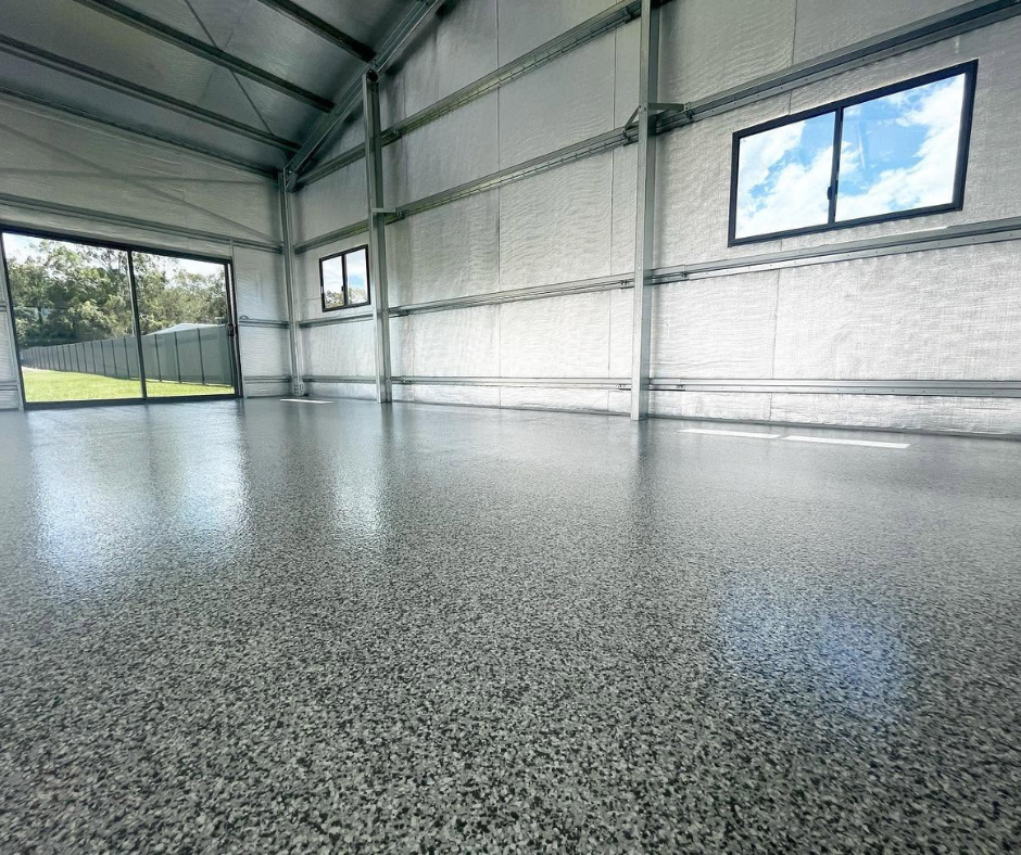 Empty industrial warehouse interior with gray speckled floor, metal walls, windows, and overhead beams