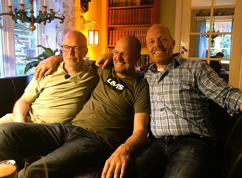 Three men on a couch, arms around each other, smiling. Interior setting with books and window.