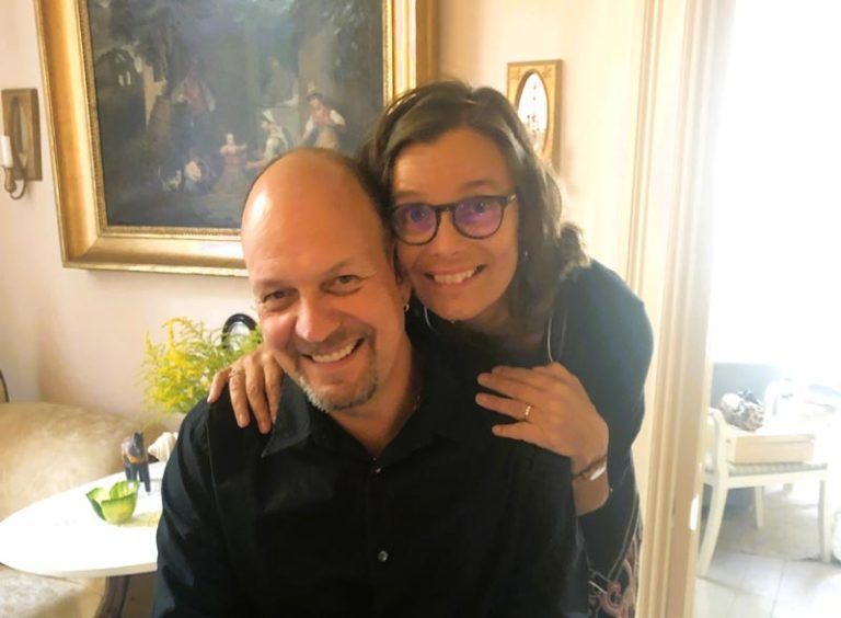 Smiling couple, woman with glasses, arm around man, posing indoors near a painting.