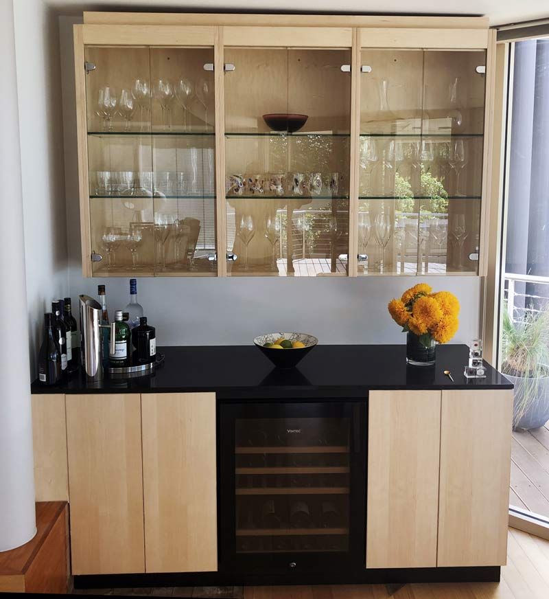 A home bar with light wood cabinets, black countertops, a wine fridge, and glass-front upper storage — Norrland in Warners Bay, NSW