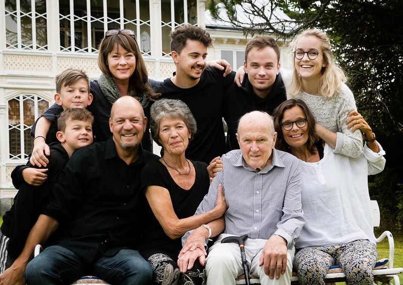 Family posing outside a house. Nine people, smiling. The elderly man sits with his cane.