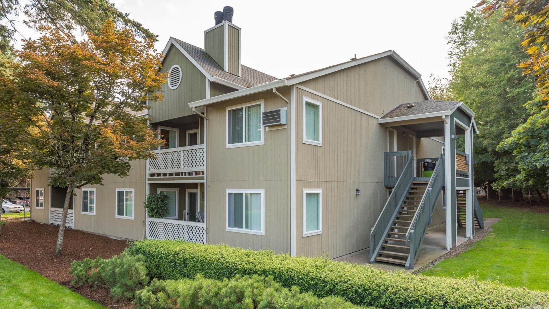 A two-story tan apartment building with a gray staircase, balconies, and green trees in a residential setting.
