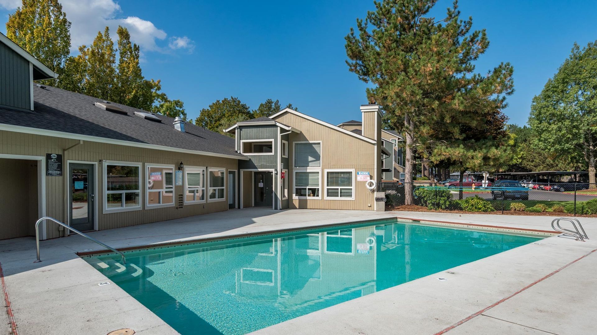 A clear swimming pool in front of a tan apartment complex under a bright blue sky on a sunny day.