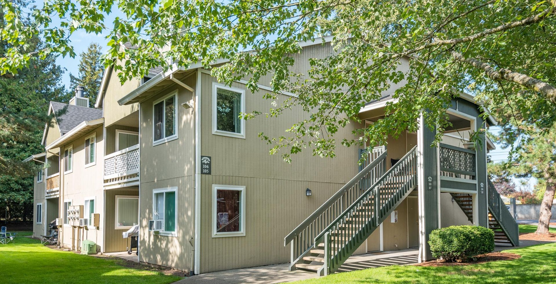 A light tan two-story apartment building with an exterior staircase, surrounded by green trees and grass on a sunny day.