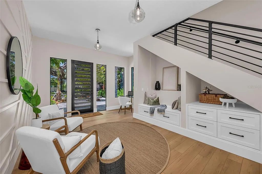 Bright modern living room with white chairs, wood floors, large windows, and a staircase railing above the built-in cabinets
