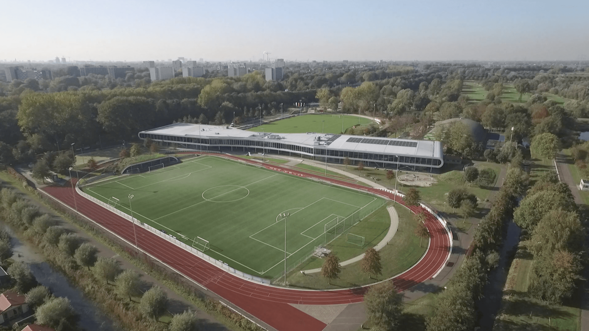 Luchtfoto van een groen sportveld met een rode atletiekbaan, omgeven door bomen en gebouwen.