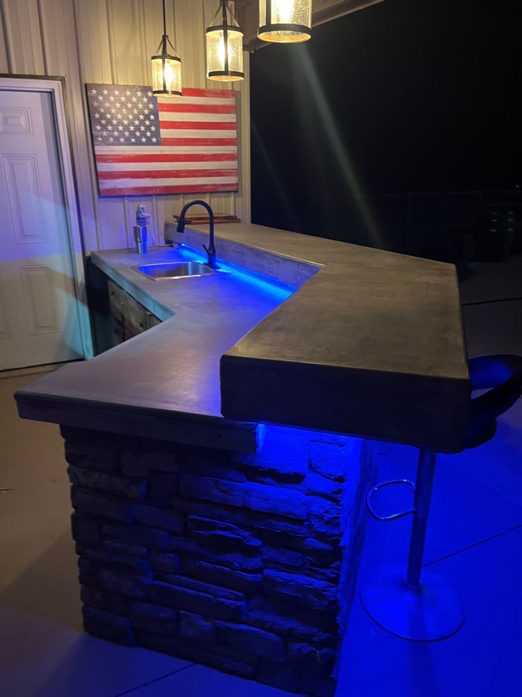 An L-shaped concrete bar with stacked stone siding, accented by blue LED lighting in an outdoor setting with an American flag