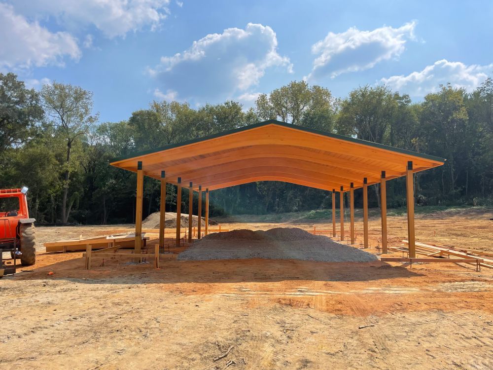 A wooden open-sided shed with a curved roof stands on a dirt construction site with a forest in the background.