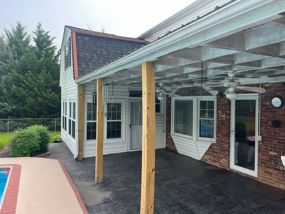 A patio area with a covered porch featuring wooden posts, adjacent to a pool deck, white siding, and a brick exterior wall.