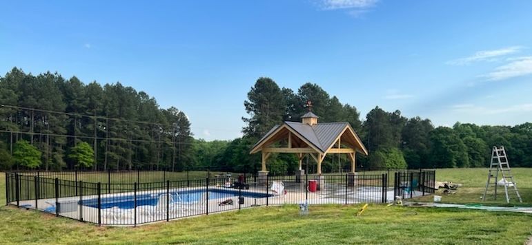 An outdoor swimming pool with a wooden pavilion, surrounded by a black metal fence, in a grassy field near a forest.