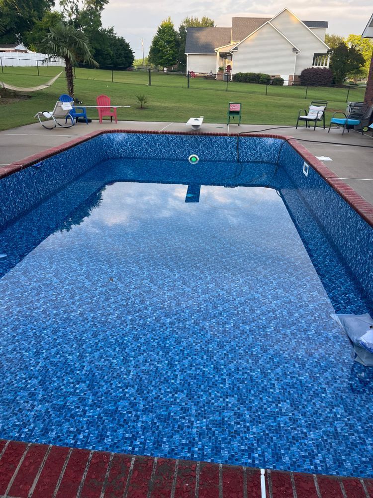 A rectangular swimming pool with blue mosaic tiles sits in a backyard on a sunny day with patio furniture and a house.