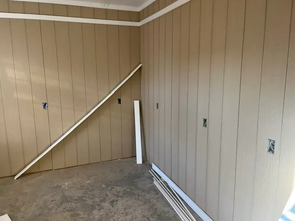 Tan vertical wall paneling in a room with white trim, electrical outlets, and loose pieces of molding on the floor.