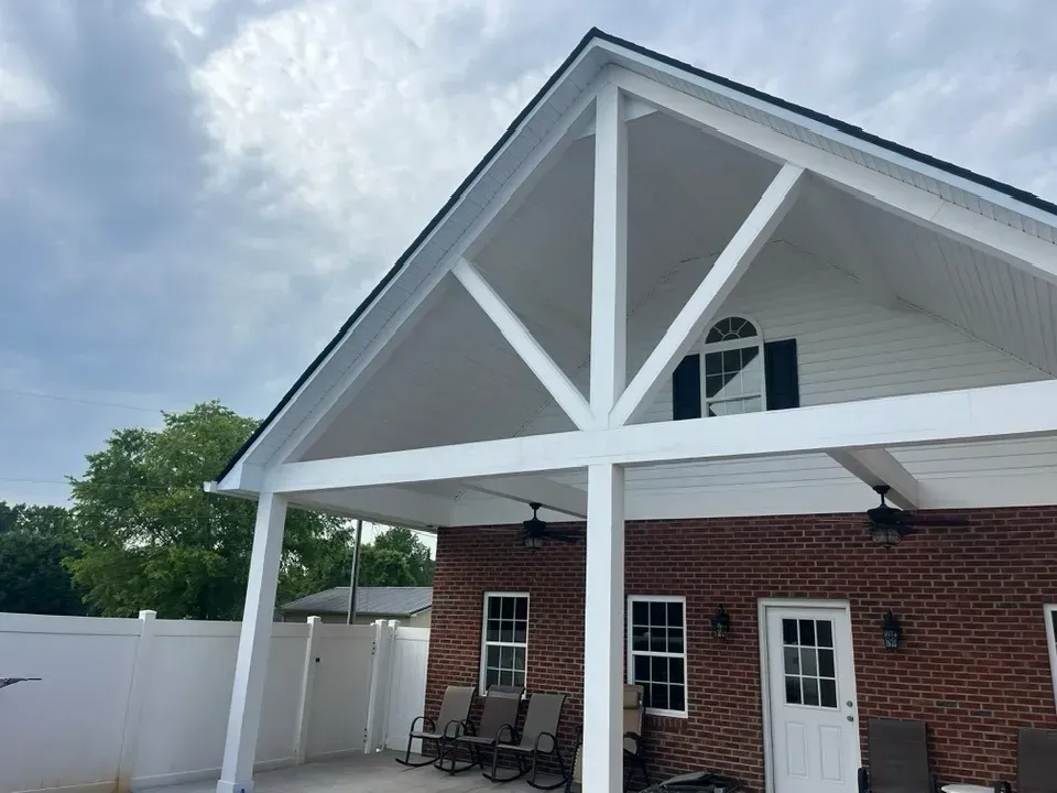 A large, white wooden patio cover over a brick house patio with outdoor furniture, a white fence, and a green tree.