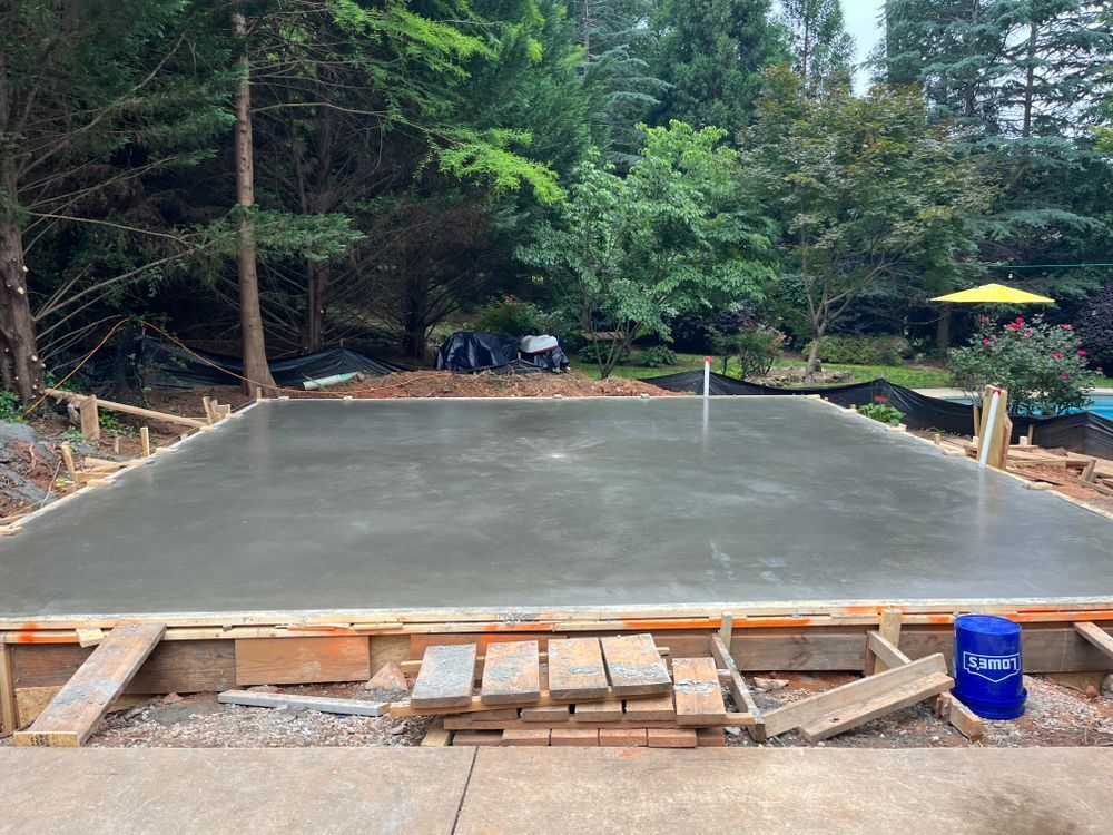 A freshly poured concrete slab foundation sits within wooden formwork in a backyard setting surrounded by trees.