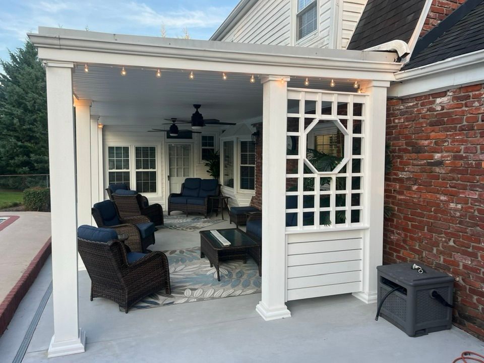 A covered patio with white pillars and a lattice screen, featuring outdoor wicker furniture arranged on a patterned rug.