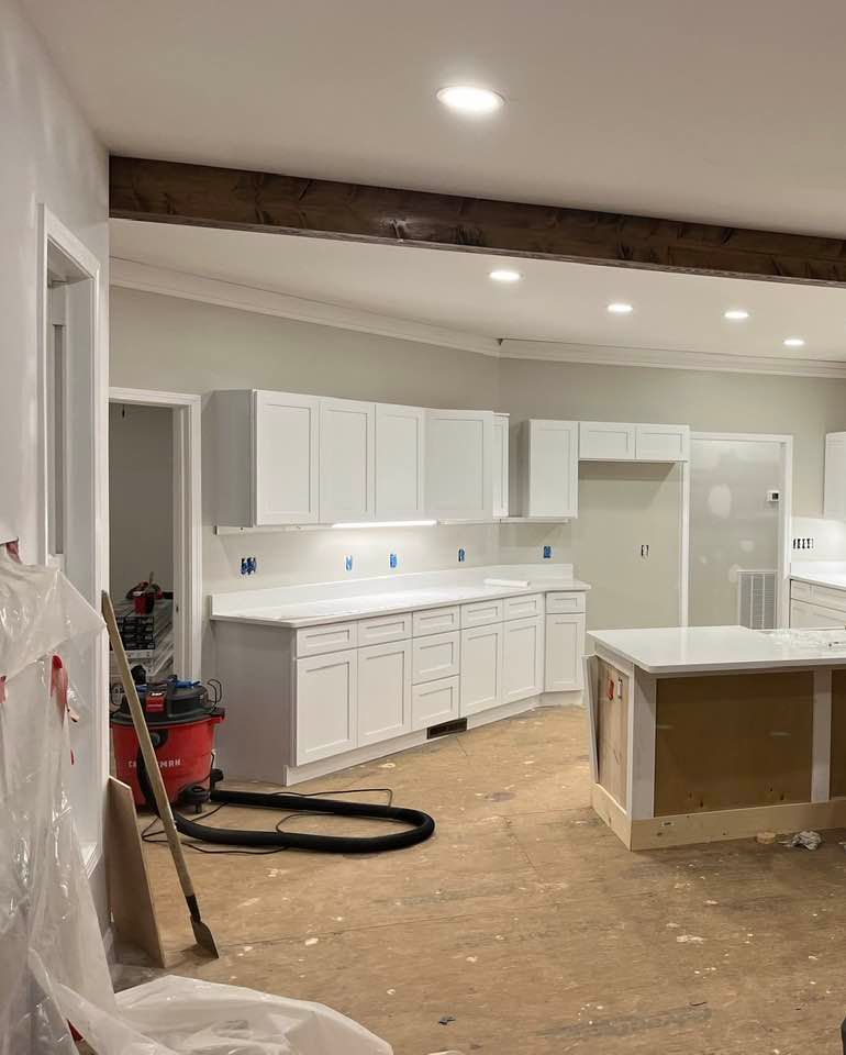A partially finished kitchen with white cabinets, white countertops, a dark wooden ceiling beam, and unfinished flooring.