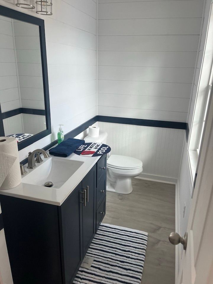 A modern bathroom with white shiplap walls, a dark navy vanity with a white countertop, and a toilet on gray tile floors.