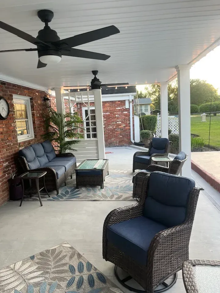 A covered brick patio with a sofa, two chairs, a coffee table, and ceiling fans, overlooking a grassy yard at sunset.