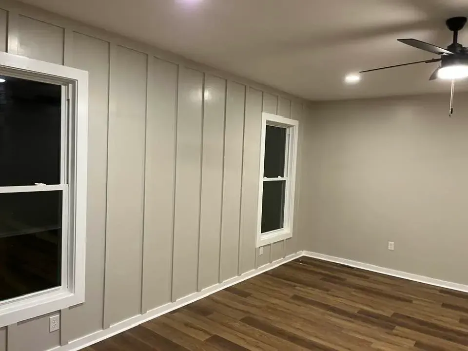 An interior room with beige board-and-batten accent wall, wood-look flooring, two windows, and a ceiling fan.