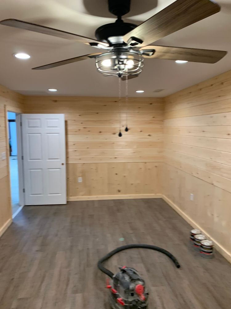 A room with wood-paneled walls, gray flooring, a ceiling fan, a white door, and a vacuum on the floor.