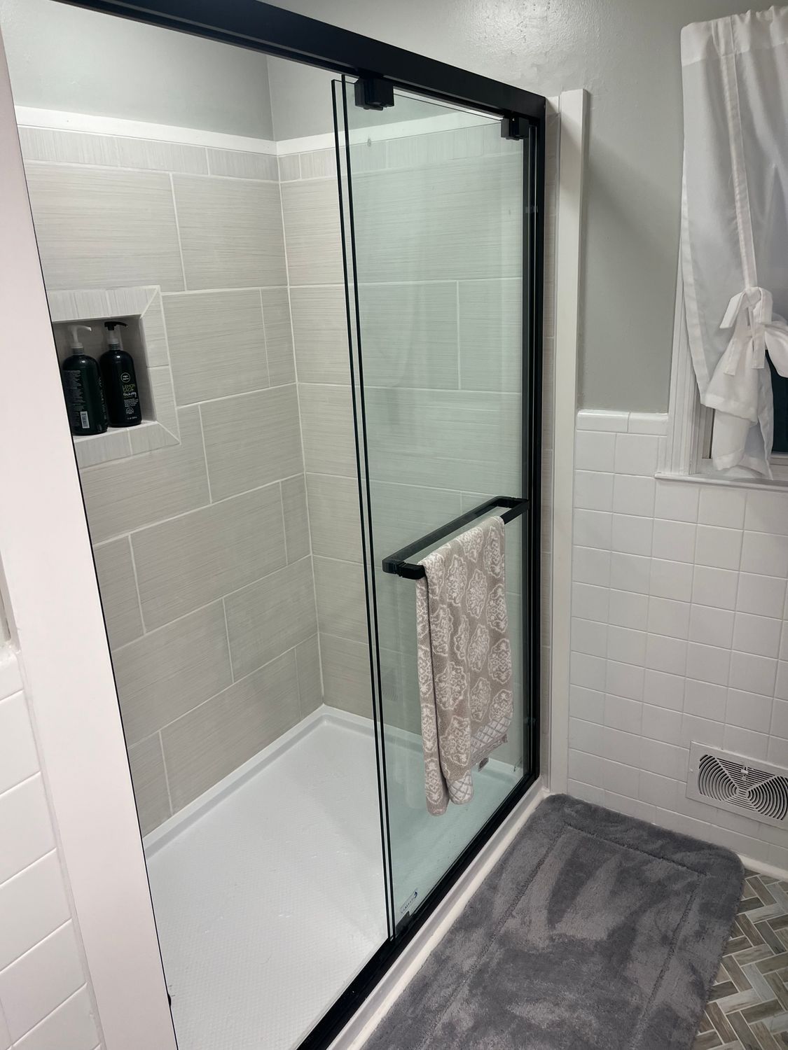 A walk-in shower with a black frame and glass sliding door, set against light-colored tiled walls and a grey floor mat.