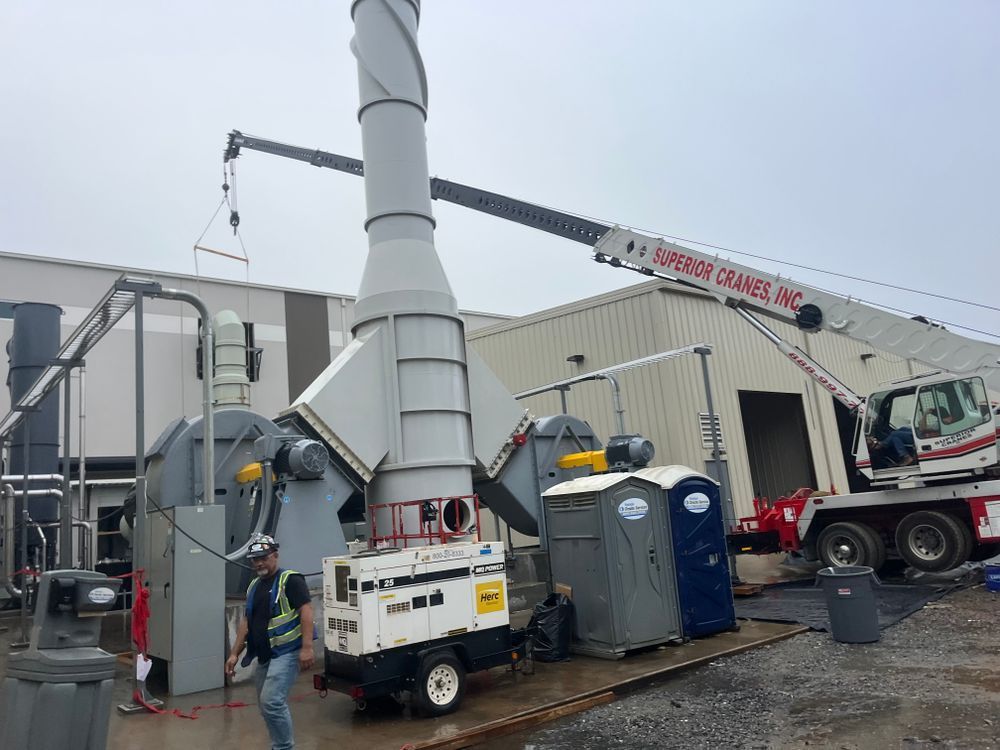 A crane lifts a large industrial exhaust stack next to factory equipment, with portable toilets on a gravel site.