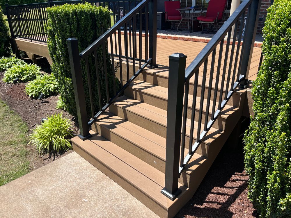 A set of tan composite stairs with black metal railings leading up to a wooden deck with greenery on the sides.