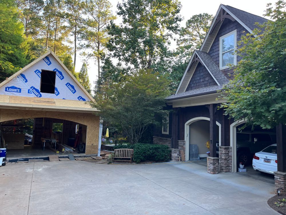 A partially constructed wooden structure stands beside a finished house with a dark brown exterior and stone-based columns.