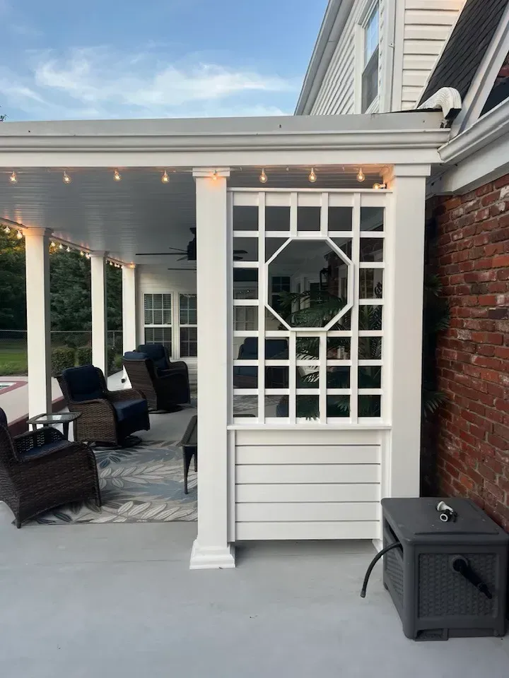 A white covered patio with a decorative trellis screen, patio furniture, and a gray hose reel on a brick home exterior.