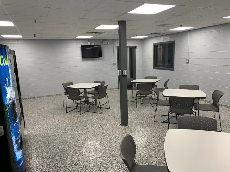 A breakroom with gray walls and speckled floors, featuring multiple small square tables with gray chairs & a vending machine.