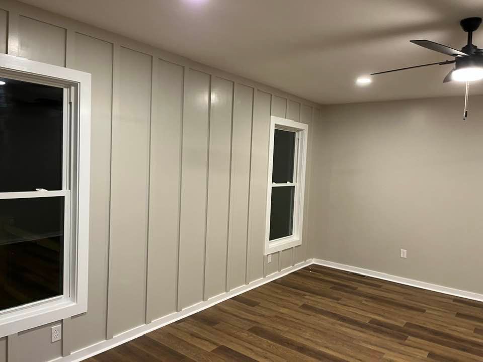 An interior room with board and batten walls, light gray paint, white trim around two windows, and wood-look flooring.