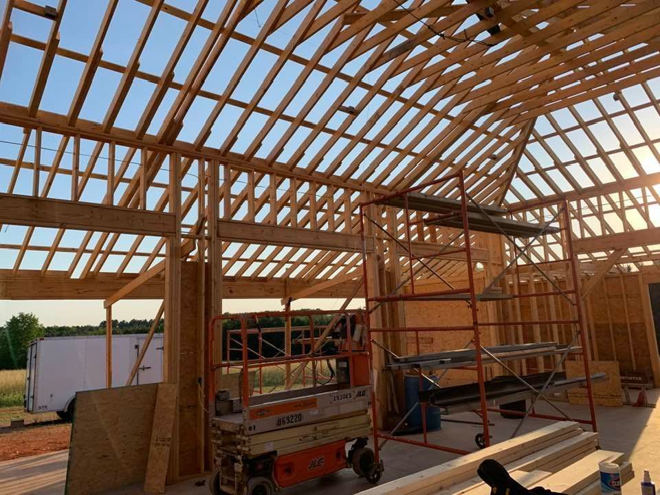 The interior framing of a wooden building under construction, featuring roof trusses, scaffolding, and a scissor lift.