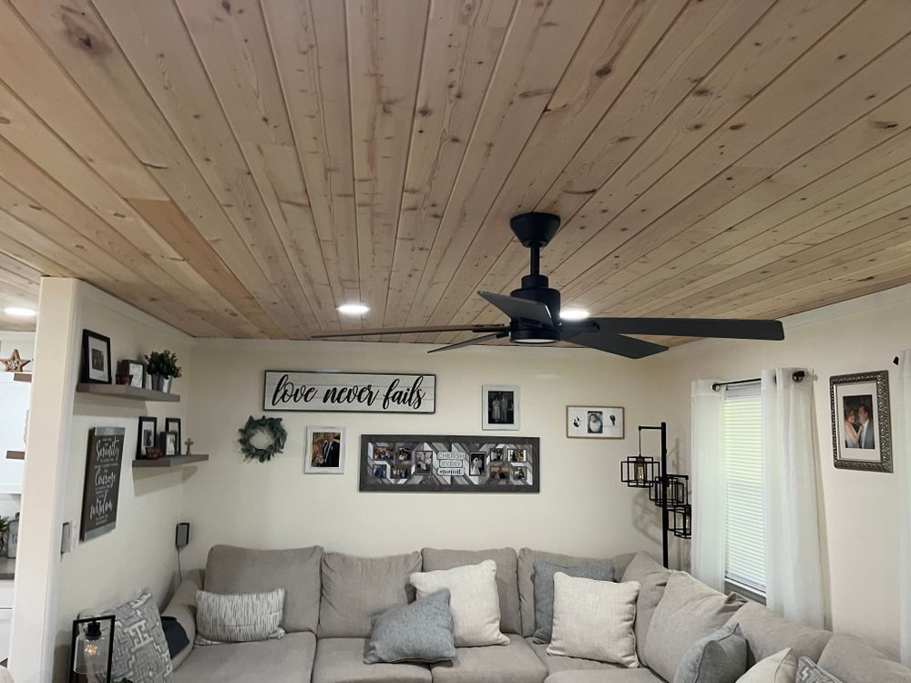 A cozy living room with a wood-plank ceiling, black ceiling fan, sectional sofa, and a wall collage of framed photos.