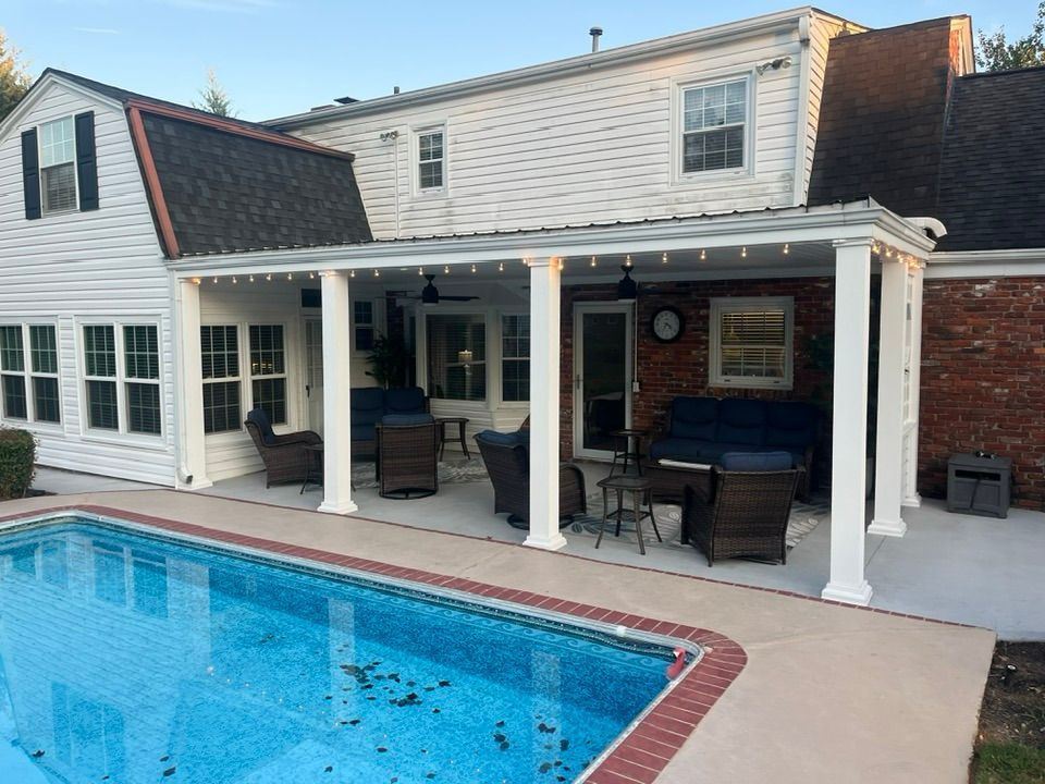 A white house with a covered patio, outdoor furniture, and string lights overlooking a backyard swimming pool.