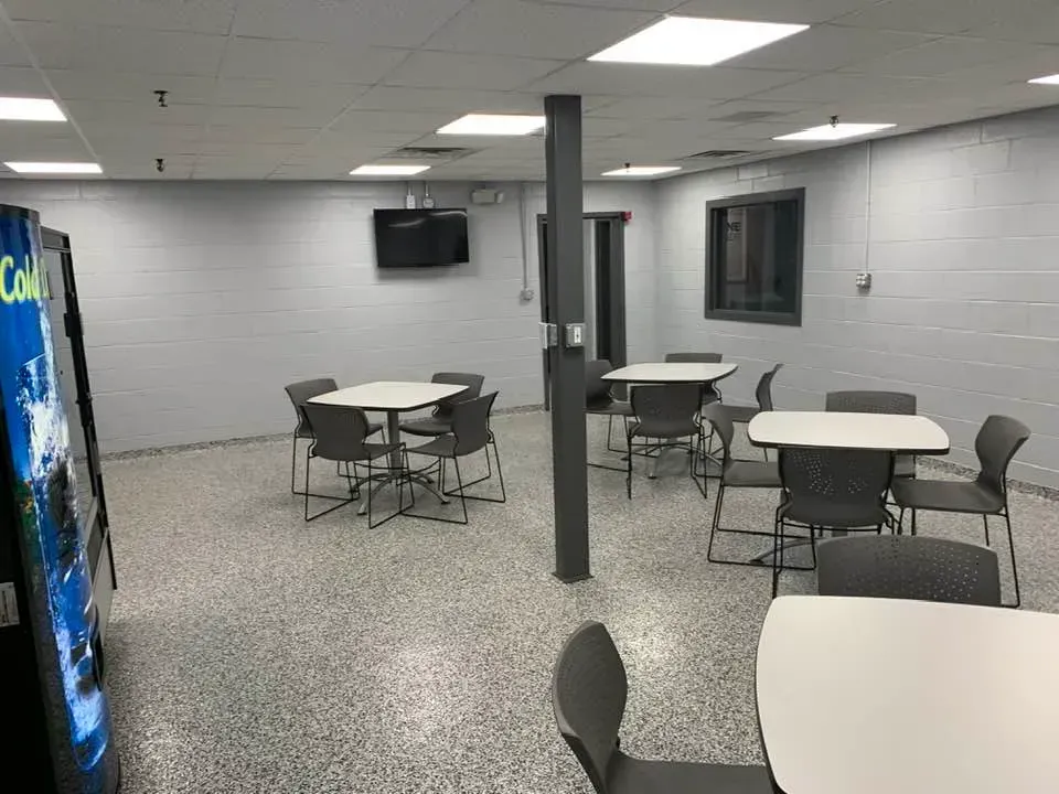 Breakroom with light gray walls, white tables, gray chairs, and a vending machine on a light gray speckled floor.
