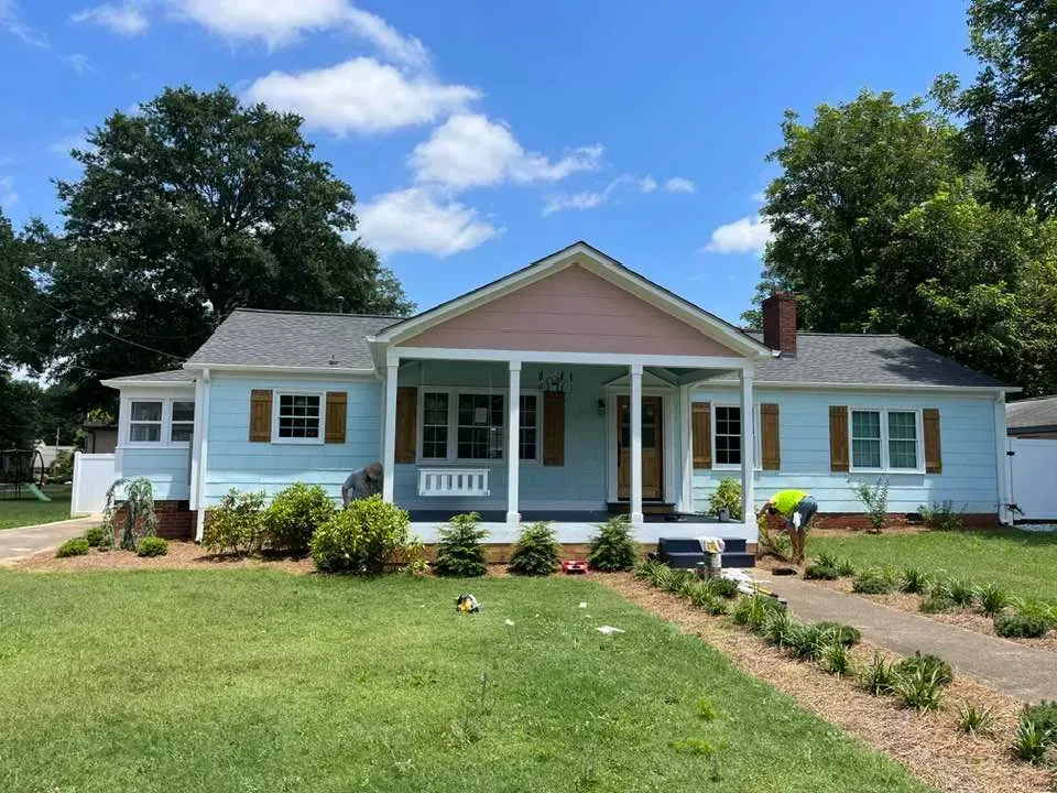 A light blue, single-story house with a white front porch and a worker landscaping the front yard under a blue sky.