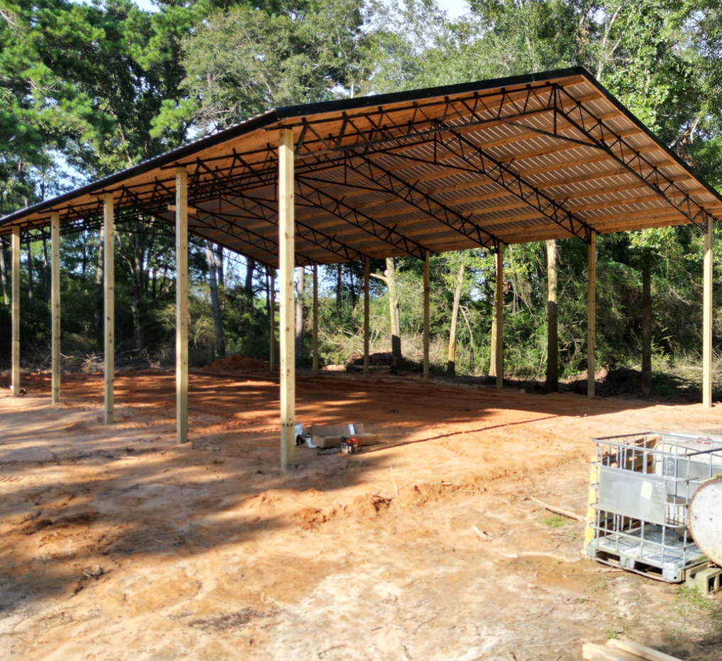 A large open-air pole barn structure with a metal roof and wooden support posts stands on a dirt lot surrounded by trees.