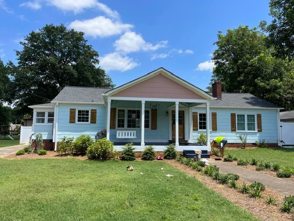 A light blue, single-story house with a white front porch, dark shutters, and a worker landscaping in the yard.
