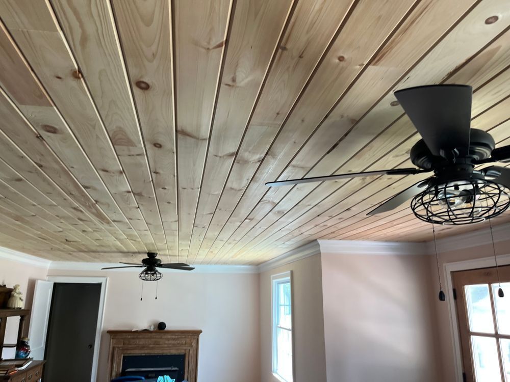 A room with a wood-plank ceiling, two black industrial ceiling fans, and light-colored walls surrounding a fireplace.