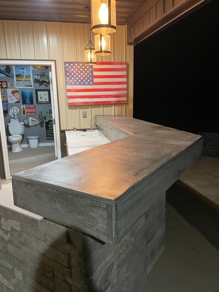 A concrete L-shaped bar with a stacked stone base sits in a room with a US flag wall hanging and pendant lights.