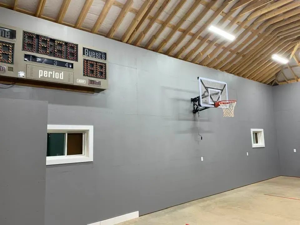 An indoor basketball court with a grey wall, a mounted hoop, and a digital scoreboard displaying game info.
