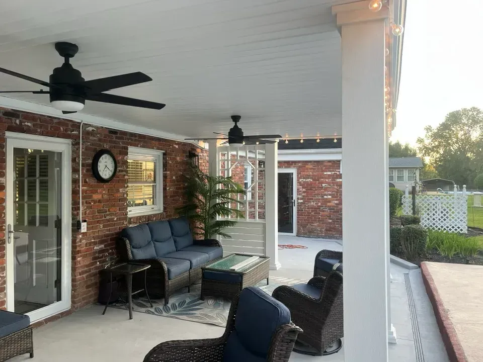 A covered brick patio furnished with a couch, armchairs, and coffee table, featuring two ceiling fans and white columns.