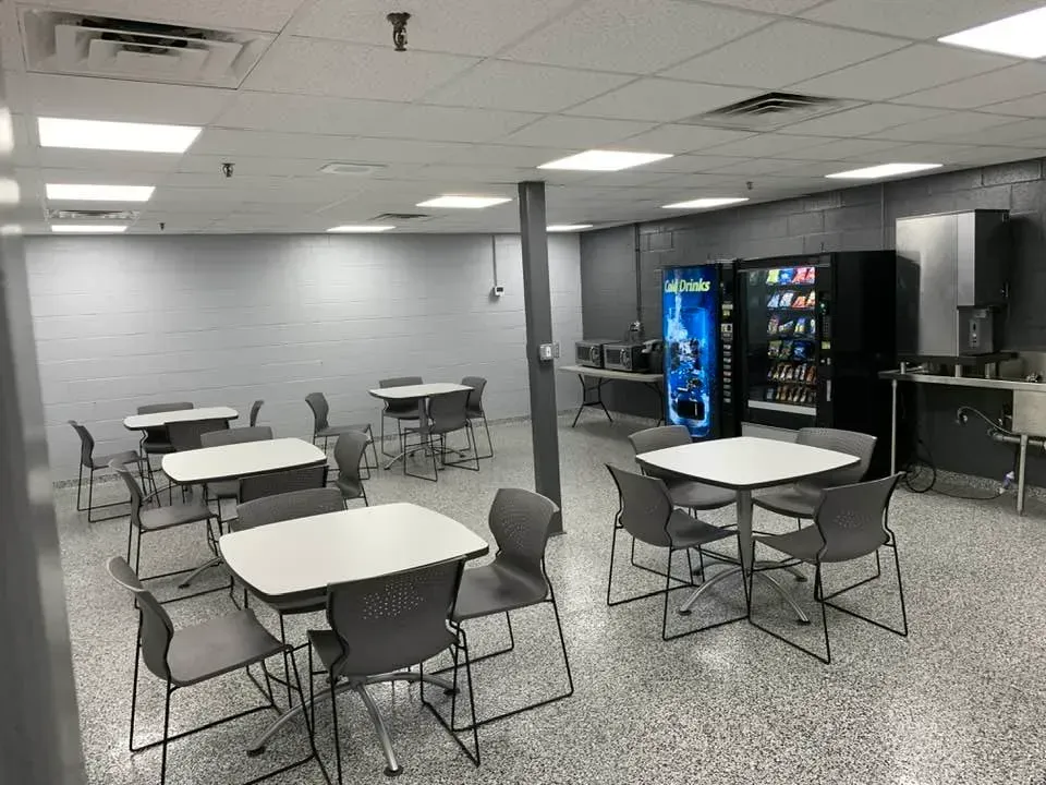 An empty breakroom with gray walls, a speckled floor, multiple tables and chairs, and two vending machines.