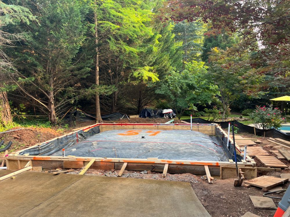 A rectangular area prepared for a concrete foundation, featuring wooden formwork and a gravel base in a wooded yard.