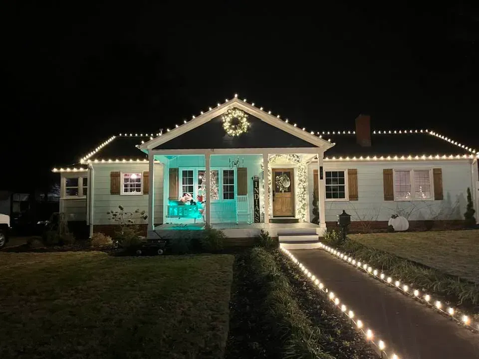 A light-blue house at night, decorated with string lights along the roof and a path lined with illuminated lanterns.