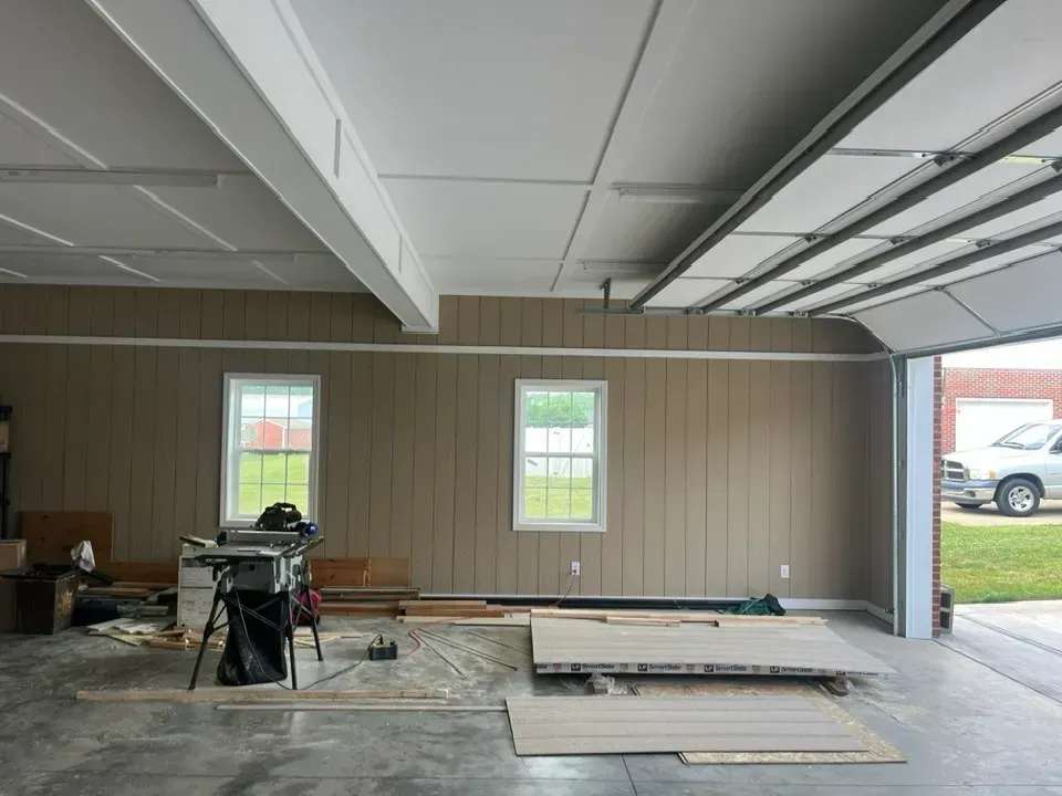 A light-filled garage workshop interior featuring two windows, a table saw, and wooden boards on the concrete floor.