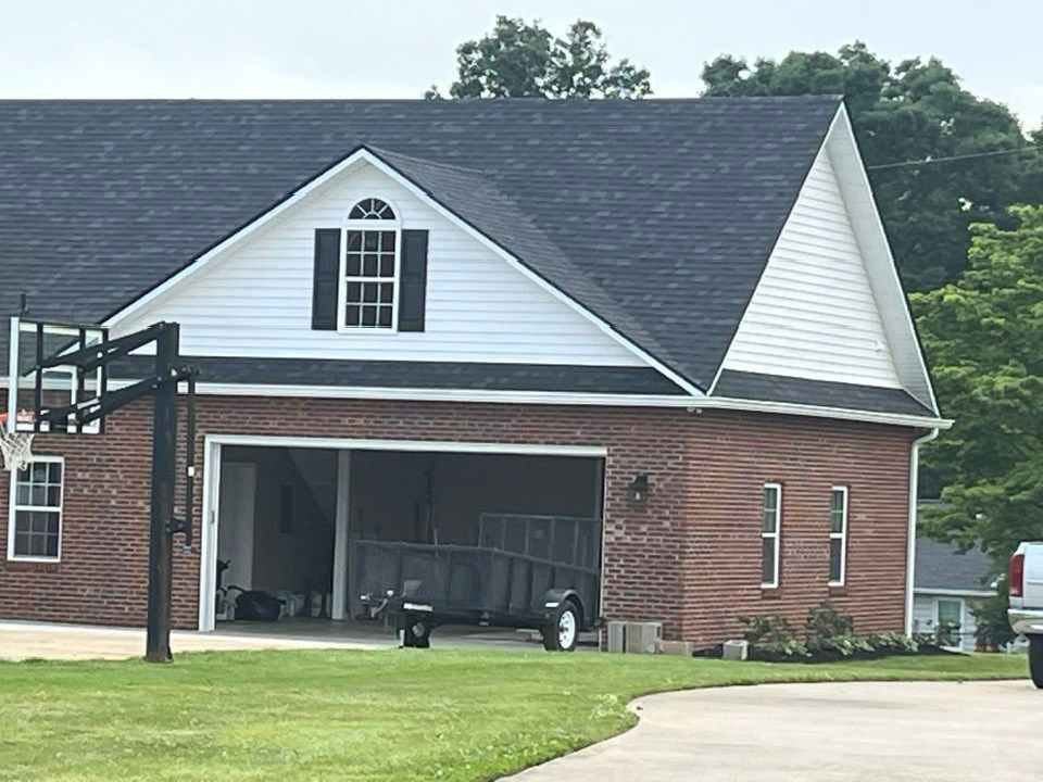 A brick garage with white siding, a peaked roof, and an open bay containing a utility trailer, with a basketball hoop.